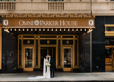 Two people in formal wedding attire stand in front of the ornate gold‑and‑black entrance of the Omni Parker House hotel. The bride’s long white veil trails across the ground as they pose beneath the illuminated sign reading “Omni Parker House,” framed by decorative lights and three arched doorways.