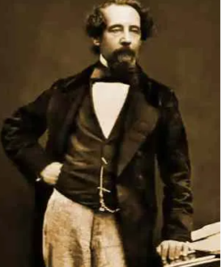   
Sepia‑toned 19th‑century portrait of a bearded man in formal Victorian attire, standing beside a small stack of books or papers. He rests one hand on the stack while the other sits on his hip, posing confidently against a plain studio backdrop.