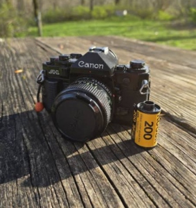  “Vintage Canon film camera on a weathered wooden surface with a roll of Kodak 200 film beside it, set outdoors near grass and trees.”