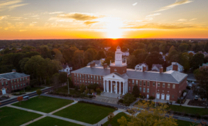 A stately brick campus building with white columns and a central clock tower stands surrounded by green lawns and trees. The sun is setting behind it, casting a golden glow across the sky and creating long, warm shadows over the grounds. Smaller brick buildings sit nearby, completing the peaceful university scene.