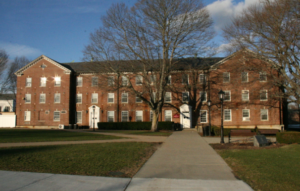 A large three‑story brick campus building with white trim and rows of windows stands behind a paved walkway. Leafless trees frame the scene, and sunlight reflects off several windows. The surrounding grass and bare branches suggest late fall or winter.