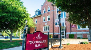   
A red‑brick multi‑story building identified as Woodward Hall at Bridgewater State University. A large maroon sign in the foreground displays the building’s name and address. Tall windows, dormers, trees, and landscaped greenery surround the hall, all lit by bright sunshine.