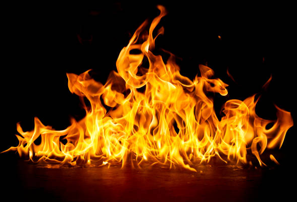   
Bright orange and yellow flames rise and curl against a completely black background. The fire burns on a flat surface, its shapes shifting and glowing sharply in the darkness.