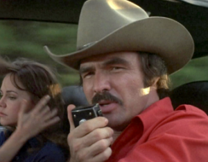   
A person in a tan cowboy hat and red shirt sits in the front seat of a moving vehicle, speaking into a CB radio microphone. Behind them, another person with long brown hair looks out the window while brushing their hair back. The blurred scenery outside suggests the vehicle is in motion.