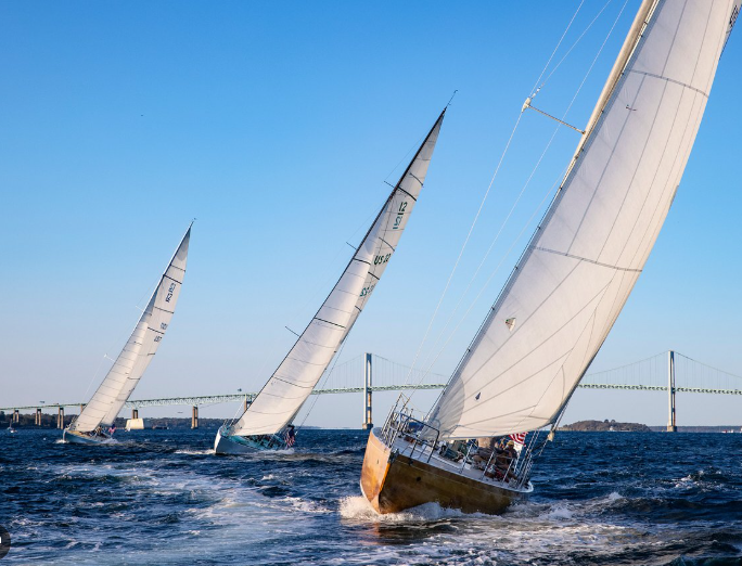 Three sailboats race across choppy blue water, each sharply heeling in the wind with sails fully extended. A long bridge stretches across the background above a low strip of land, and the sky is bright and clear. The scene captures the speed and intensity of the America’s Cup in Newport.