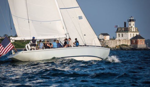  A white sailboat with full sails moves through choppy water, carrying several people and an American flag at the stern. A lighthouse and a few shoreline buildings sit on a rocky point in the background, capturing the classic coastal setting of the America’s Cup in Newport.