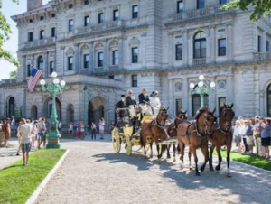 A horse‑drawn carriage with four brown horses approaches an ornate stone mansion as a crowd looks on. People in formal or period clothing ride in the yellow carriage, and an American flag hangs near the building’s entrance. The scene takes place on a sunny day with trees casting partial shade.