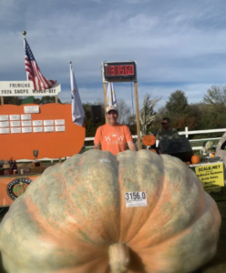 A man stands behind an enormous pumpkin weighing 3,156 pounds at an outdoor weigh‑off event. The pumpkin sits on a platform with a digital display showing its weight, and an orange event board with flags is visible in the background.