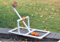 A small homemade catapult built from white PVC pipes sits on a concrete surface outdoors. The angled launching arm holds an orange ball in a plastic cup, with two more orange balls resting nearby. Fallen leaves scatter across the grassy background, showing a simple DIY setup for launching small objects.