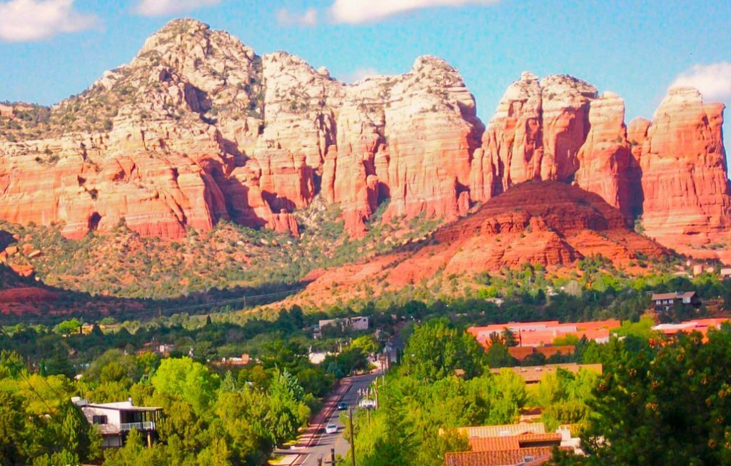  Red rock formations rise above the town of Sedona, Arizona, with layered sandstone cliffs, green trees, and scattered houses under a clear sky.