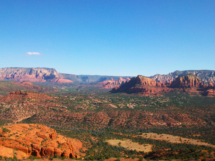 Wide desert landscape with red rock mesas and buttes in Sedona, Arizona, surrounded by sparse vegetation and green shrubs under a clear blue sky.