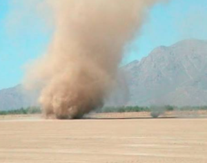  A tall dust devil spirals upward from the desert floor, twisting across a flat, dry landscape with distant mountains under a clear blue sky.