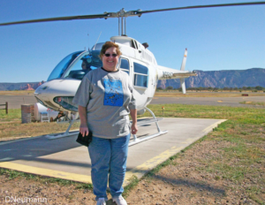 A woman stands in front of a white sightseeing helicopter on a desert helipad, wearing sunglasses, a graphic T‑shirt, and jeans, with rocky hills and a clear blue sky in the background during her first helicopter ride.