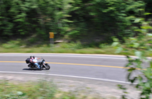   
A motorcycle with two riders leans into a curve on a forested road, motion blur showing their speed as they pass dense green foliage and a yellow road sign.
