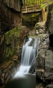   
A narrow waterfall flows between tall moss‑covered rock walls in a shaded gorge, with a wooden footbridge above and green forest vegetation surrounding the scene.