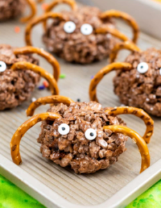 Chocolate-covered rice cereal treats decorated as playful spiders, with pretzel-stick legs and candy eyes, arranged on a light baking tray with colorful sprinkles scattered around. Bright lighting highlights the crunchy texture and whimsical Halloween-themed design.
