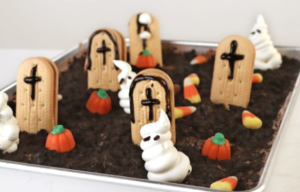 Halloween-themed dessert decorated to look like a graveyard. A metal baking pan is filled with dark chocolate crumb topping resembling soil, with cookie tombstones decorated with black crosses standing throughout the dessert. White ghost-shaped frosting figures, small orange pumpkin candies, and pieces of candy corn are scattered across the surface. The dessert is photographed from an angled overhead view against a light background.