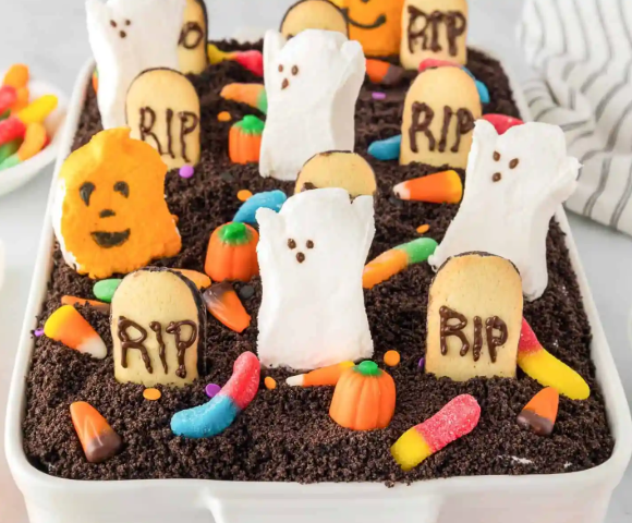 Halloween-themed graveyard dessert in a white baking dish, topped with crushed chocolate cookie "dirt," marshmallow ghosts, cookie tombstones decorated with "RIP" in chocolate, candy corn, gummy worms, and small pumpkin candies. An orange ghost-shaped cookie with a jack-o'-lantern face stands near the front, while additional ghost and tombstone decorations fill the dessert, creating a colorful and playful spooky scene.