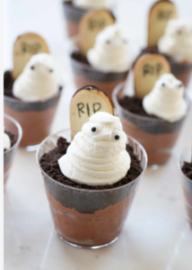 alloween-themed chocolate pudding cups topped with crushed cookie "dirt," white ghost-shaped whipped cream decorations with candy eyes, and small tombstone cookies labeled "RIP." Several dessert cups are arranged on a light-colored surface, creating a playful spooky cemetery effect.
