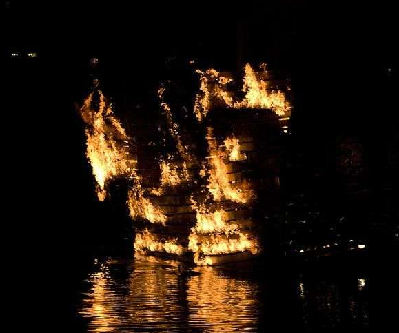 A burning ship at night, flames and reflections on the water evoking the legend of the Palatine Light.