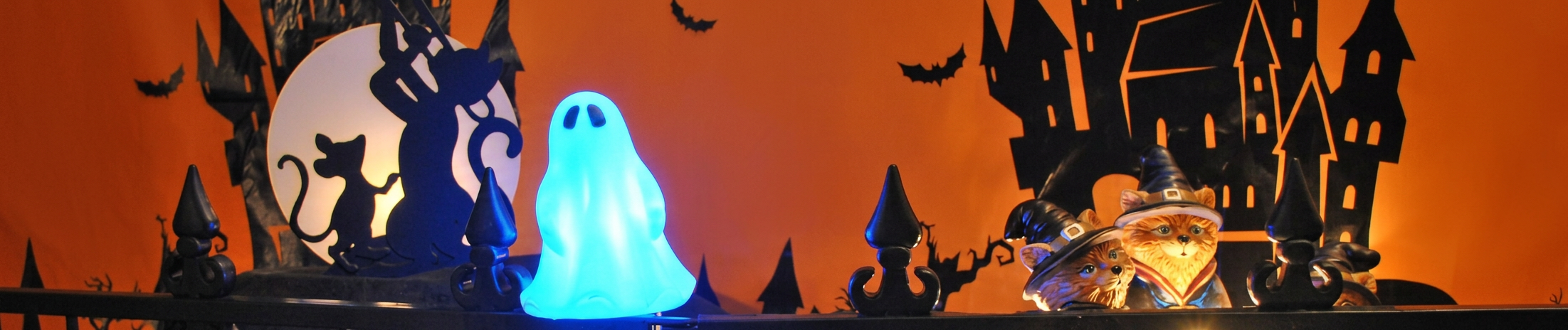 Halloween-themed banner featuring an orange background with black silhouettes of haunted houses, bats, and a full moon. In the foreground, a glowing blue ghost decoration stands beside two orange tabby cat figurines wearing witch hats. Black decorative fence posts frame the scene, creating a festive Halloween display with warm orange lighting and spooky accents.