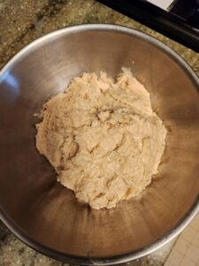  A stainless steel mixing bowl holds a rough, partially mixed ball of dough resting on a speckled kitchen countertop.