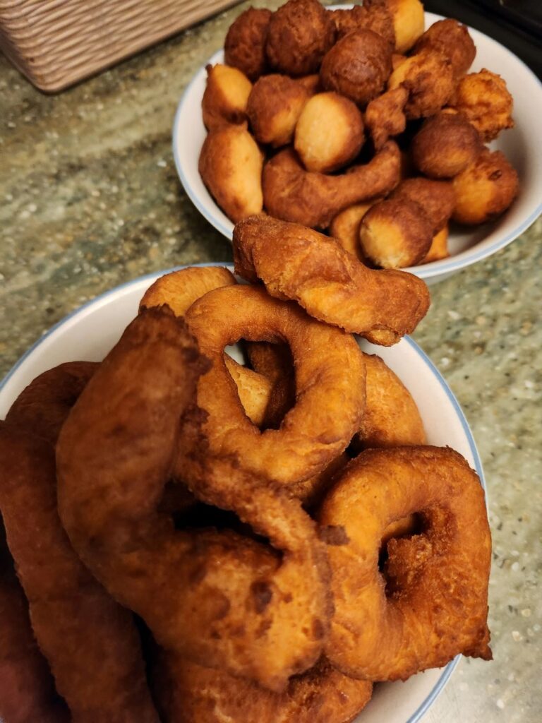 Two bowls of freshly fried donuts sit on a kitchen counter—one filled with large ring‑shaped donuts, the other with smaller round and oval donut holes, all golden brown and crisp.