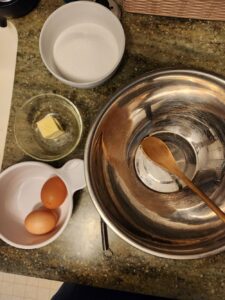  A kitchen countertop with a large metal mixing bowl and wooden spoon, plus small bowls holding butter, eggs, and granulated sugar arranged for baking.