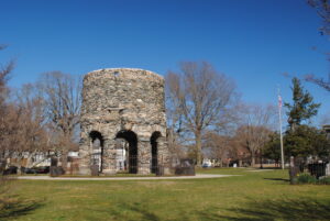 The Newport Tower stands in a grassy park, its round stone structure supported by several archways. Leafless trees surround the site, with a few buildings and a flagpole visible in the background under a bright blue sky.